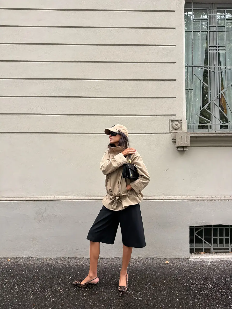 A beige belted jacket and baseball cap styled with black Bermuda shorts, sunglasses, a black shoulder bag and leopard-print slingback shoes.