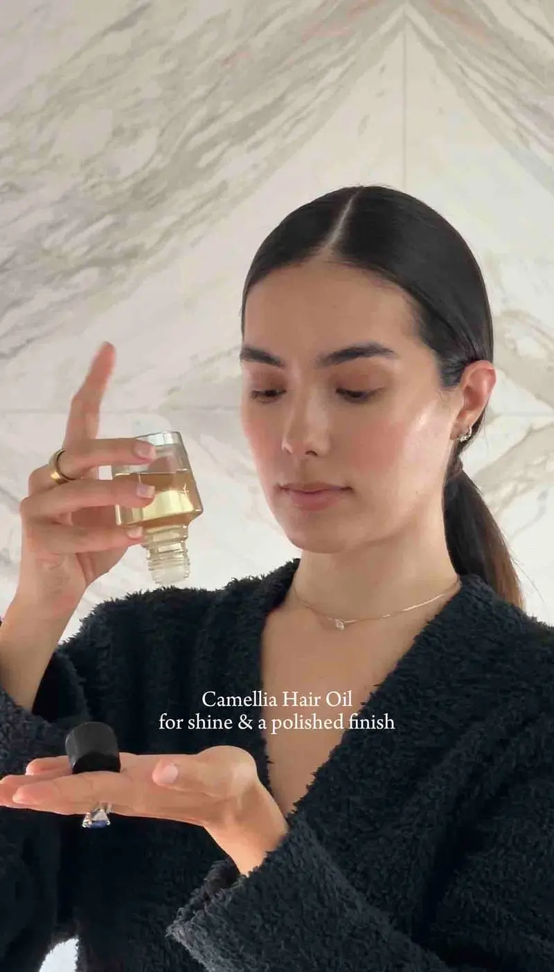 Sharon with a sleek, center-parted low ponytail, pouring hair oil into her palm while wearing a black textured top.