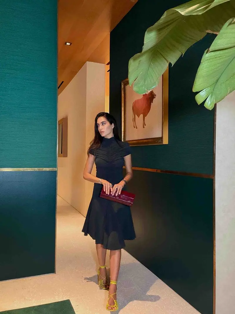 Sharon wearing a dark short-sleeved dress, bright yellow-green strappy heels, and a burgundy clutch in a room with green walls.