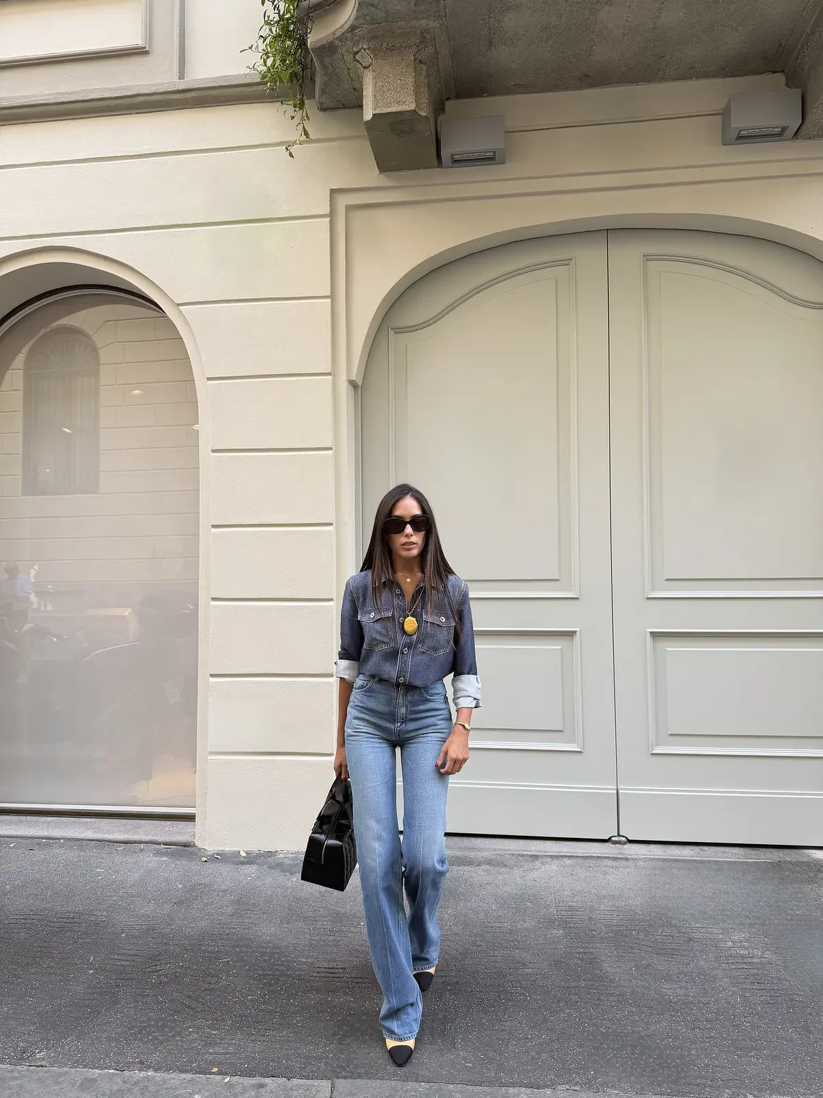 Sharon con camisa vaquera, jeans azules, zapatos bicolores y bolso negro frente a una puerta en Milán.