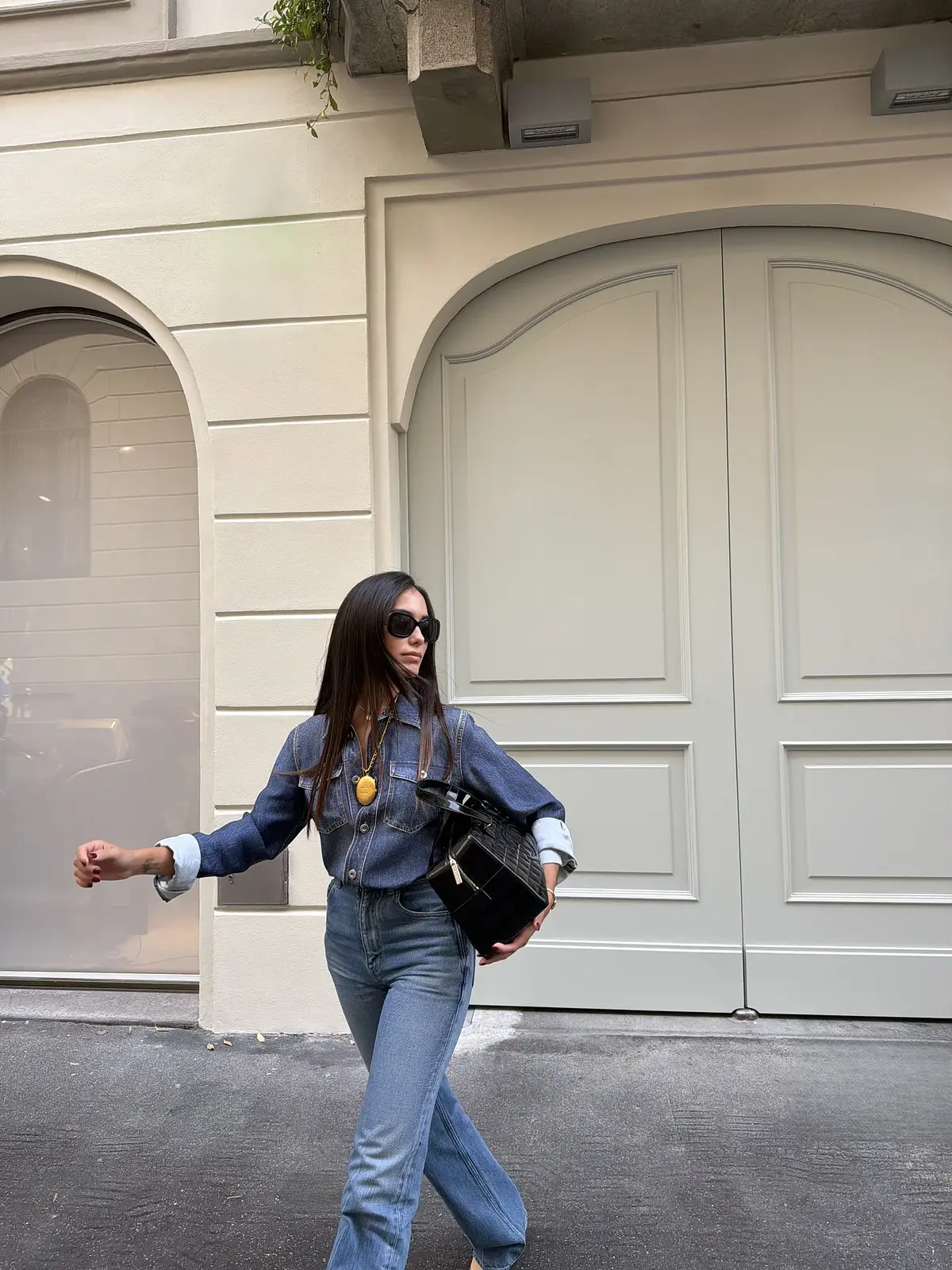 Sharon cammina con camicia in denim, jeans, occhiali da sole e una borsa nera sotto il braccio.